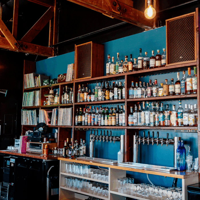 A wall of the back of a bar. There are three shelves lined up on the wall of various alcohol. There are tap handles located below.