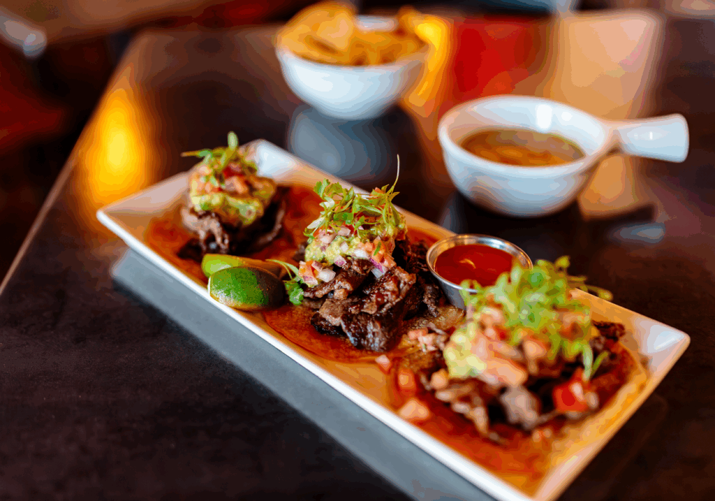 A long white plate of three beef tacos. In the background is are a bowl of chips and bowl of salsa.