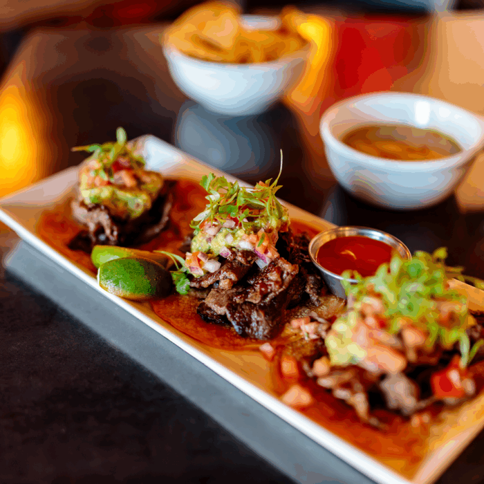 A long white plate of three beef tacos. In the background is are a bowl of chips and bowl of salsa.